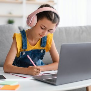 Schoolgirl in pink headset sitting at desk, writing in textbook