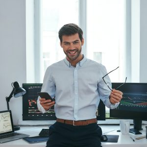 Happy young businessman or trader in formalwear holding eyeglasses and smartphone