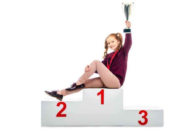 smiling schoolgirl sitting on winner podium, holding trophy cup in raised hand and looking at camera